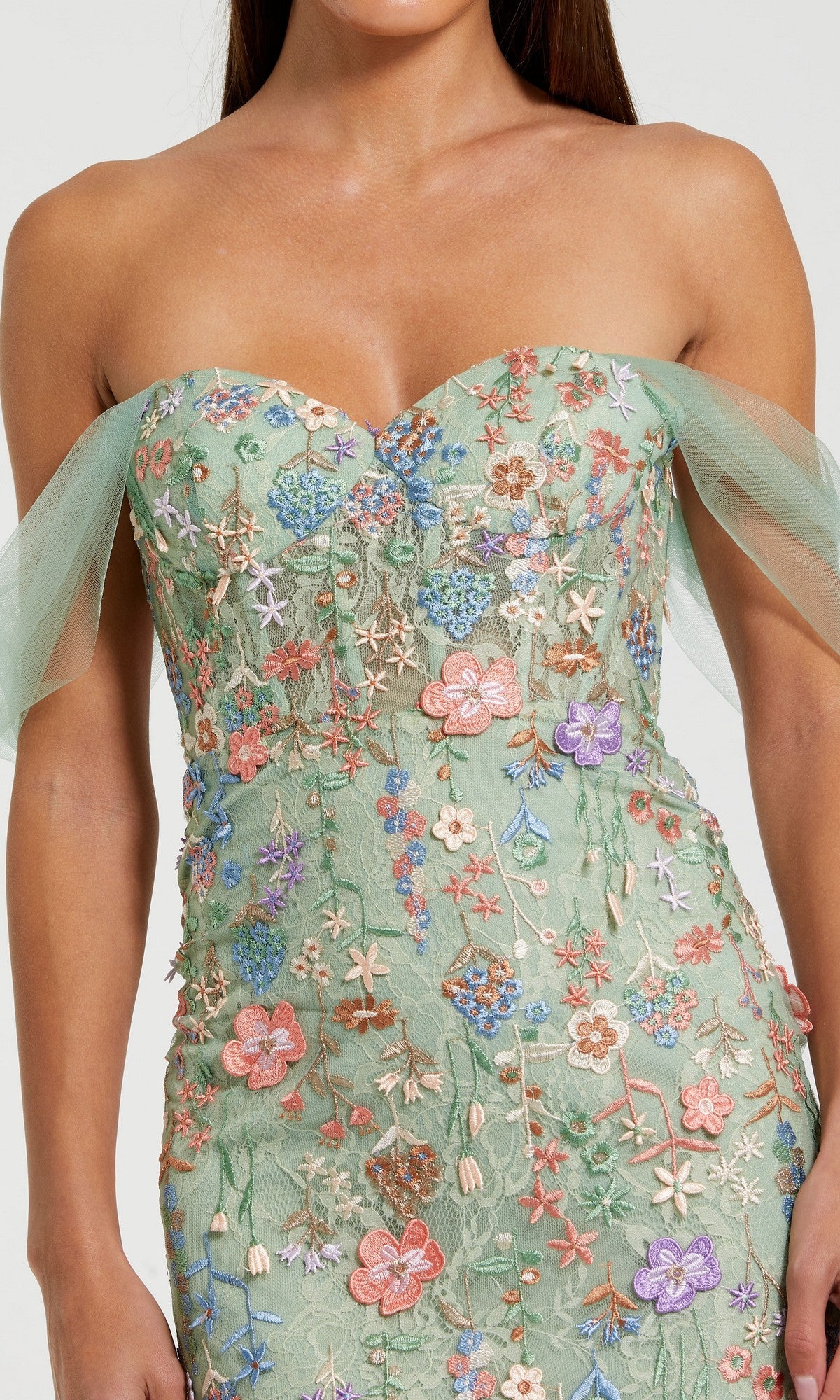 A woman wears the Mac Duggal 68809 long formal dress in light green, with an off-the-shoulder fitted corset bodice, tulle sleeves, and colorful floral embroidery.